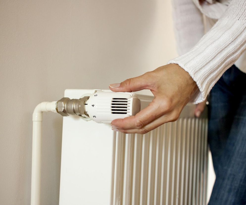 Ręka osoby w białym swetrze i dżinsach reguluje biały termostat grzejnika. Suche powietrze z ogrzewania wpływa na zdrowie dróg oddechowych, a o tym, jak temu zaradzić, przeczytasz na Poradnik Zdrowie.
