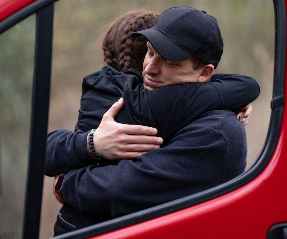 Celina (Orina Krajewska) w czarnej bluzie z warkoczem na ramieniu i Łukasz (Michał Rolnicki) w czarnej czapce z daszkiem oraz ciemnej bluzie w uścisku, widoczni przez otwarte okno czerwonego samochodu. Oboje obejmują się mocno, a twarz Łukasza wyraża ulgę lub szczęście. Więcej o ich losach dowiesz się na Super Seriale.