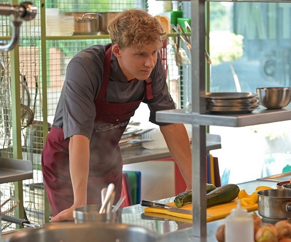 Młody kucharz Tomek w czerwonym fartuchu, stojący przy stalowym blacie kuchennym, wpatruje się w przestrzeń z zamyśloną miną. W tle widać półki z naczyniami, zieloną kratownicę i jasne okno. Obraz oddaje emocjonalne wyzwania Tomka z serialu Pierwsza miłość, o których można przeczytać na Super Seriale.