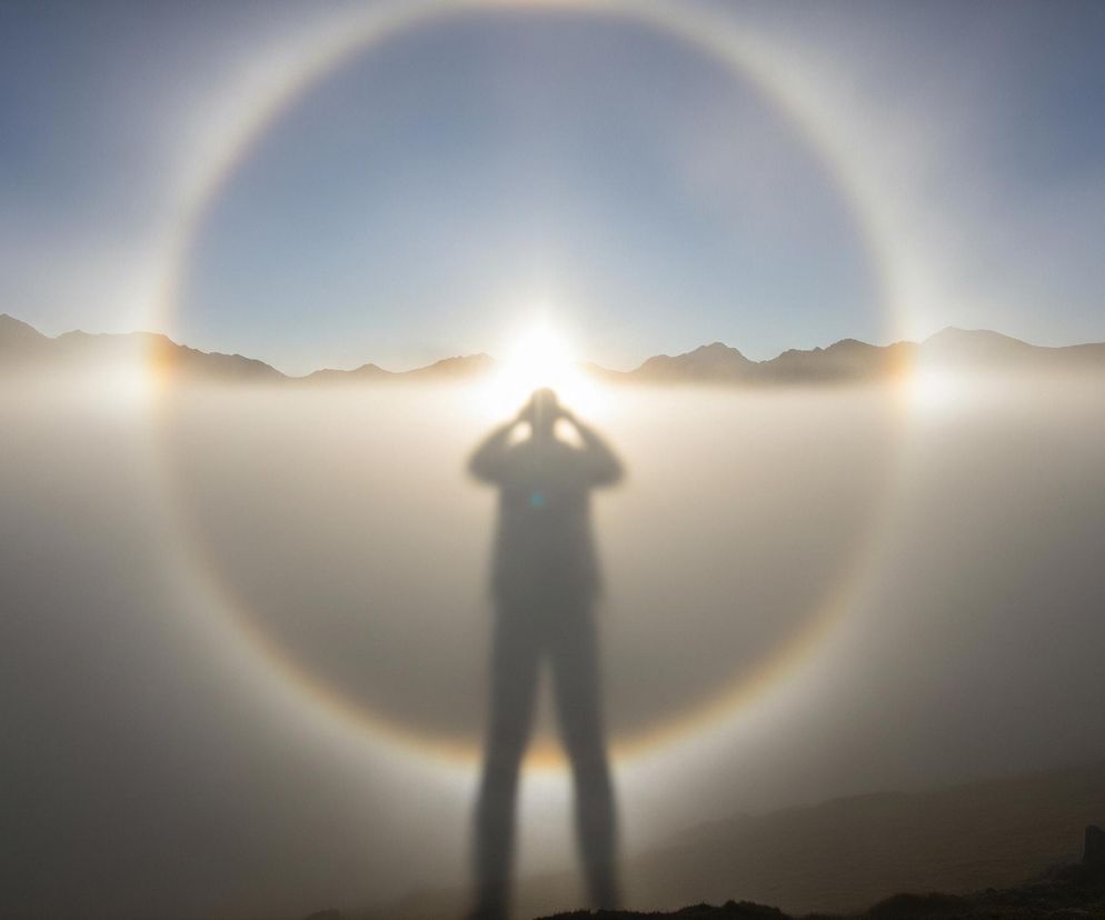 Na pierwszym planie widoczny jest ciemny, rozmyty zarys postaci człowieka, stojącego na niewyraźnym, ciemnym podłożu. Ręce postaci są uniesione, sprawiając wrażenie, jakby trzymała aparat fotograficzny. Bezpośrednio za postacią, w centralnej części kadru, znajduje się jasne słońce, otoczone wyraźnym, okrągłym halo z delikatnymi, tęczowymi barwami. Tło wypełnia warstwa mgły lub chmur, przez którą przebijają się ciemne sylwetki górskich szczytów, a powyżej rozciąga się jasnoniebieskie niebo.