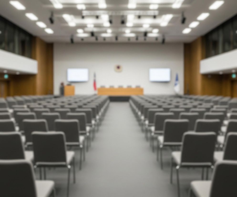 Duże pomieszczenie konferencyjne jest wypełnione rzędami pustych krzeseł z ciemnoszarym obiciem, ustawionych na szarej wykładzinie. Krzesła tworzą dwie główne sekcje, rozdzielone centralnym przejściem prowadzącym w głąb sali. Na końcu pomieszczenia znajduje się podwyższona scena z długim, drewnianym stołem i kilkoma krzesłami, a także dwa białe ekrany po obu stronach i mównica z lewej strony. Sufit jest jasny, z licznymi wpuszczanymi oprawami oświetleniowymi i widocznymi elementami nagłośnienia.