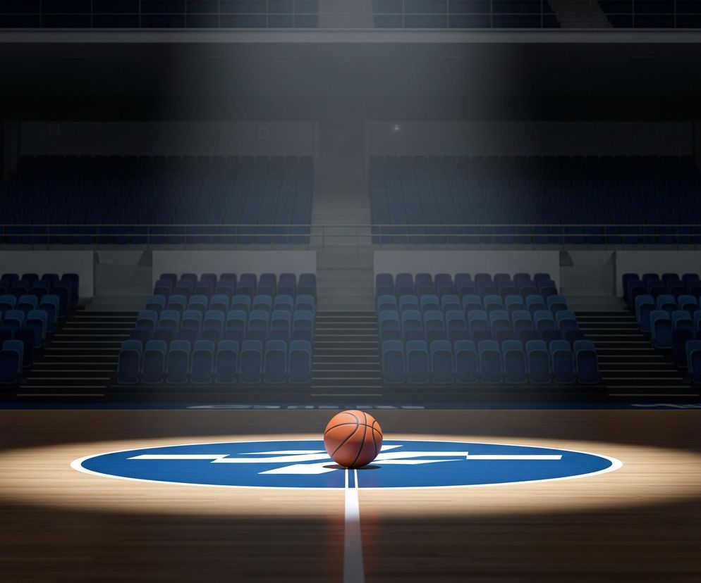 Na drewnianym parkiecie, oświetlonym silnym, okrągłym strumieniem światła, leży pomarańczowa piłka do koszykówki. Znajduje się ona na centralnym, niebieskim okręgu z białymi oznaczeniami, na linii biegnącej przez środek boiska. Tło stanowi puste audytorium z rzędami ciemnoniebieskich siedzeń, rozciągających się na trybunach pod częściowo zaciemnionym sufitem, z którego przebija kilka punktowych świateł.