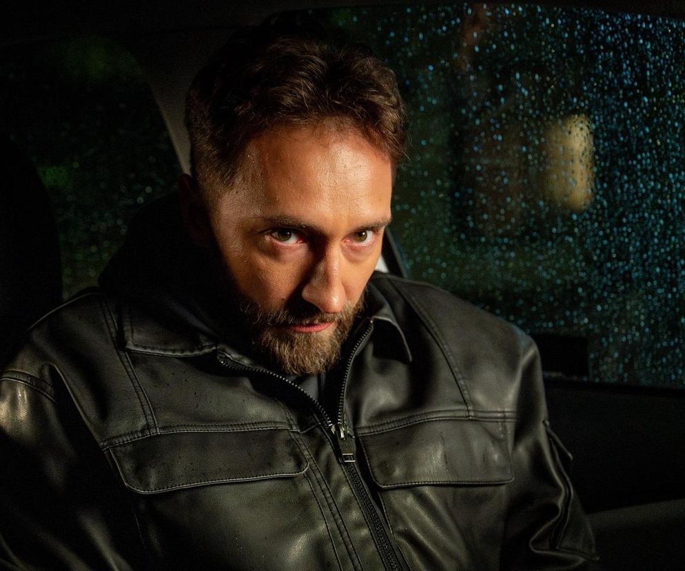 Dariusz Wieteska jako Piotr Strzelecki w skórzanej kurtce, z brodą, siedzący w samochodzie, patrzący intensywnie. Scena przedstawia niebezpieczną sytuację Piotra, wynikającą z uzależnienia od leków. Więcej o dramacie ratownika przeczytasz na Super Seriale.