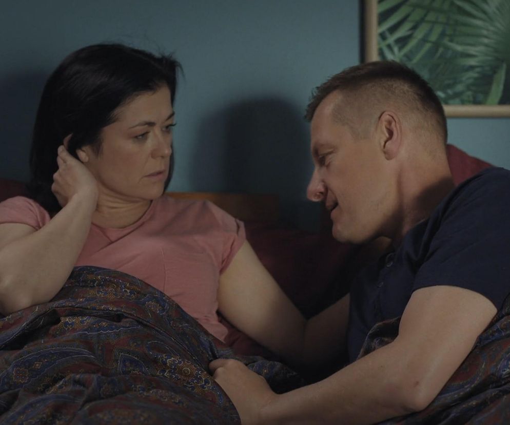 Katarzyna Cichopek (Kinga Zduńska) i Marcin Mroczek (Piotrek Zduński) w sypialni, oboje pod kołdrą z kolorowym, orientalnym wzorem, Kinga w różowej koszulce, Piotrek w granatowej. Kinga patrzy na Piotrka z zamyśleniem, trzyma rękę przy uchu. Piotrek zwraca się do niej, próbując ją pocieszyć po traumatycznych wydarzeniach. Scena z serialu, o której przeczytasz więcej na Super Seriale.