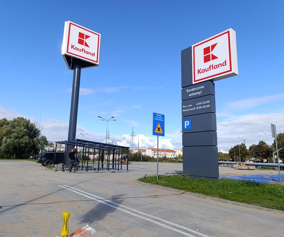Znak pionowy z logo Kauflandu oraz duży pylon informacyjny ze szczegółami otwarcia nowego sklepu w Rumi, widoczne na tle częściowo zachmurzonego nieba. Obok znaku budowana jest wiata rowerowa. Więcej o konflikcie Kauflandu z OPZZ na Super Biznes.