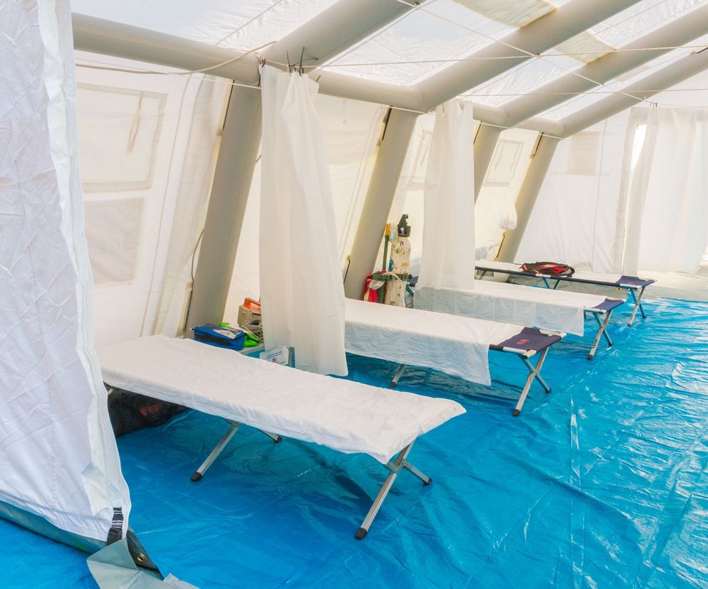 Wnętrze namiotu szpitala polowego z rzędem pustych, prowizorycznych łóżek polowych pokrytych białym materiałem, ustawionych na niebieskiej plandece. Widoczne są elementy konstrukcji namiotu oraz wiszące białe zasłony, symbolizujące tymczasową infrastrukturę medyczną, o której więcej przeczytasz na Poradnik Zdrowie.