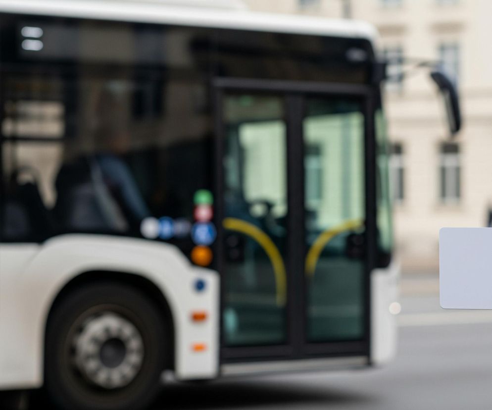 Prawa ręka trzyma białą, prostokątną kartę zbliżeniową przed czarnym terminalem, z niewyraźnym tłem przedstawiającym duży, biały autobus miejski z czarnymi szybami i elementami konstrukcyjnymi. Autobus stoi na ulicy, za nim widać rozmyte, beżowe budynki z oknami i sylwetki przechodniów. Karta jest ostro wykadrowana na pierwszym planie, podczas gdy pozostałe elementy obrazu są rozmyte.