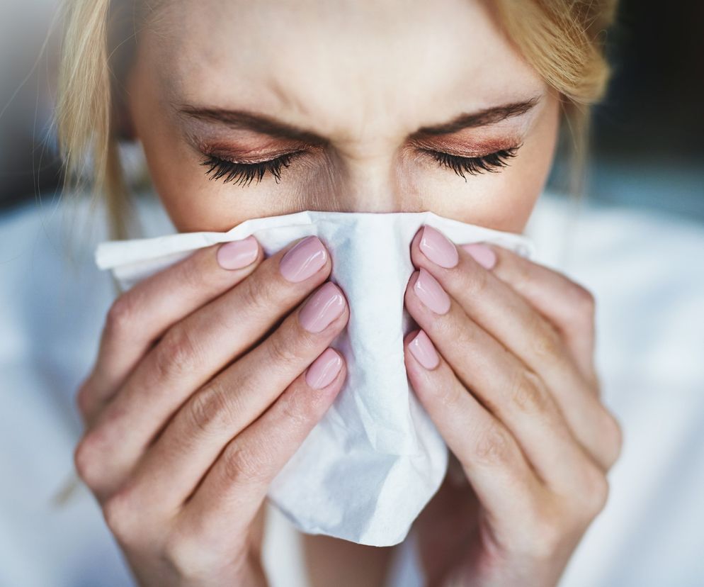 Kobieta z zamkniętymi oczami, zdmuchująca nos w białą chusteczkę, odczuwająca dyskomfort z powodu kataru, co ilustruje temat bezpiecznego leczenia przeziębienia. Dowiedz się więcej o domowych sposobach na katar i zatkany nos na Poradnik Zdrowie.