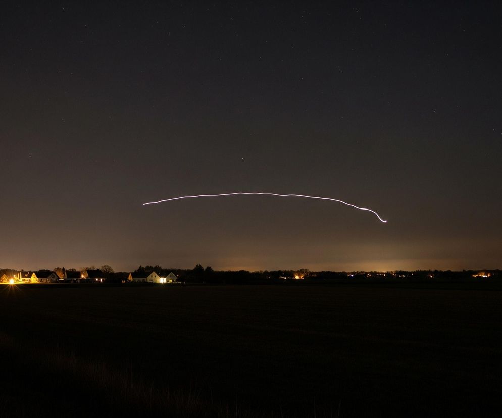Na ciemnym niebie rozciąga się cienka, falista, biało-fioletowa linia, biegnąca od lewej do prawej, delikatnie opadając w prawym końcu. Poniżej, na horyzoncie, widoczne są światła wsi lub małego miasteczka, tworzące żółtawą poświatę. Na pierwszym planie rozciąga się ciemne pole uprawne, zajmujące dolną połowę kadru, z delikatnie widocznymi zarysami roślinności.
