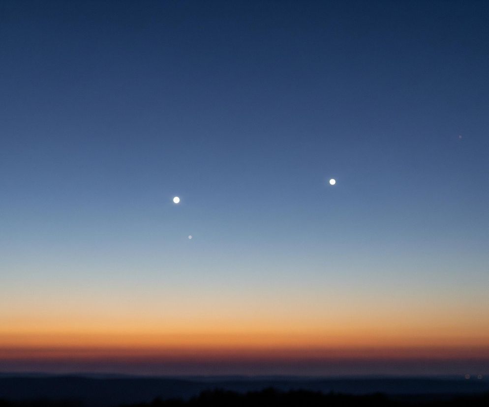 Niebo w górnej części obrazu jest ciemnoniebieskie, stopniowo jaśniejące ku horyzontowi, gdzie przechodzi w odcienie pomarańczy i żółci. Na tle nieba widoczne są trzy jasne, białe punkty, przypominające gwiazdy lub planety, ułożone w niemal poziomą linię. Poniżej pomarańczowej linii horyzontu rozciąga się ciemny, niewyraźny pejzaż, prawdopodobnie pagórkowaty teren, ze smugi jaśniejszych świateł w oddali.