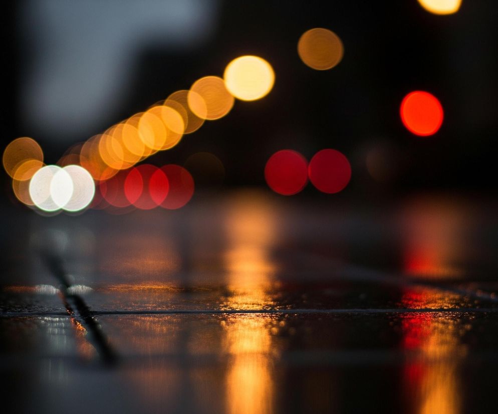 Na obrazie widoczne są rozmyte, okrągłe światła bokeh w różnych kolorach, odbijające się od mokrej, ciemnej powierzchni. Górna część kadru wypełniona jest serią pomarańczowych świateł, układających się skośnie od lewej do prawej, z kilkoma czerwonymi i białymi punktami świetlnymi. Dolna połowa zdjęcia przedstawia lśniącą, odbijającą światła nawierzchnię, prawdopodobnie asfalt lub bruk, tworząc pionowe smugi światła. Całość utrzymana jest w ciemnych barwach, z dominującymi odcieniami czerni, pomarańczu i czerwieni, a także akcentami bieli.