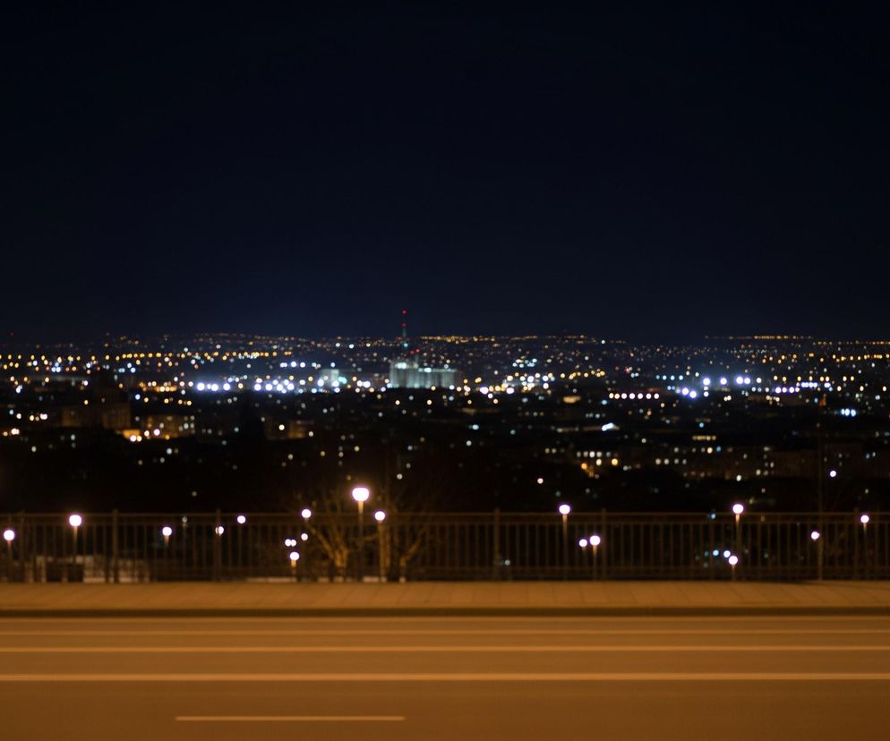 Nocna panorama miasta rozświetlonego licznymi światłami, widziana z podwyższonej perspektywy. Na pierwszym planie znajduje się szeroka, asfaltowa droga z białymi liniami, a za nią widoczny jest chodnik i niski, metalowy płot. Wzdłuż płotu rozmieszczone są uliczne latarnie o kulistych, jasnych kloszach, rzucające żółte światło. W tle rozciąga się miejska zabudowa, pełna rozproszonych, jasnych punktów świetlnych, które tworzą efekt bokeh na ciemnym niebie.