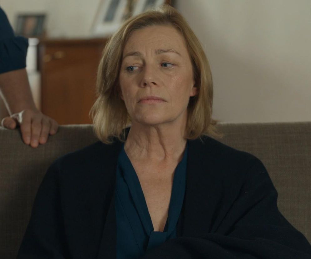 Maja Barełkowska jako Wanda siedzi na kanapie w ciemnym ubraniu, z zamyśloną miną, a zza niej wyłania się dłoń Tomasza (Jakub Sokołowski) w granatowej koszuli, dotykająca oparcia. Scena z odcinka „Barwy szczęścia dostępnego na Super Seriale.