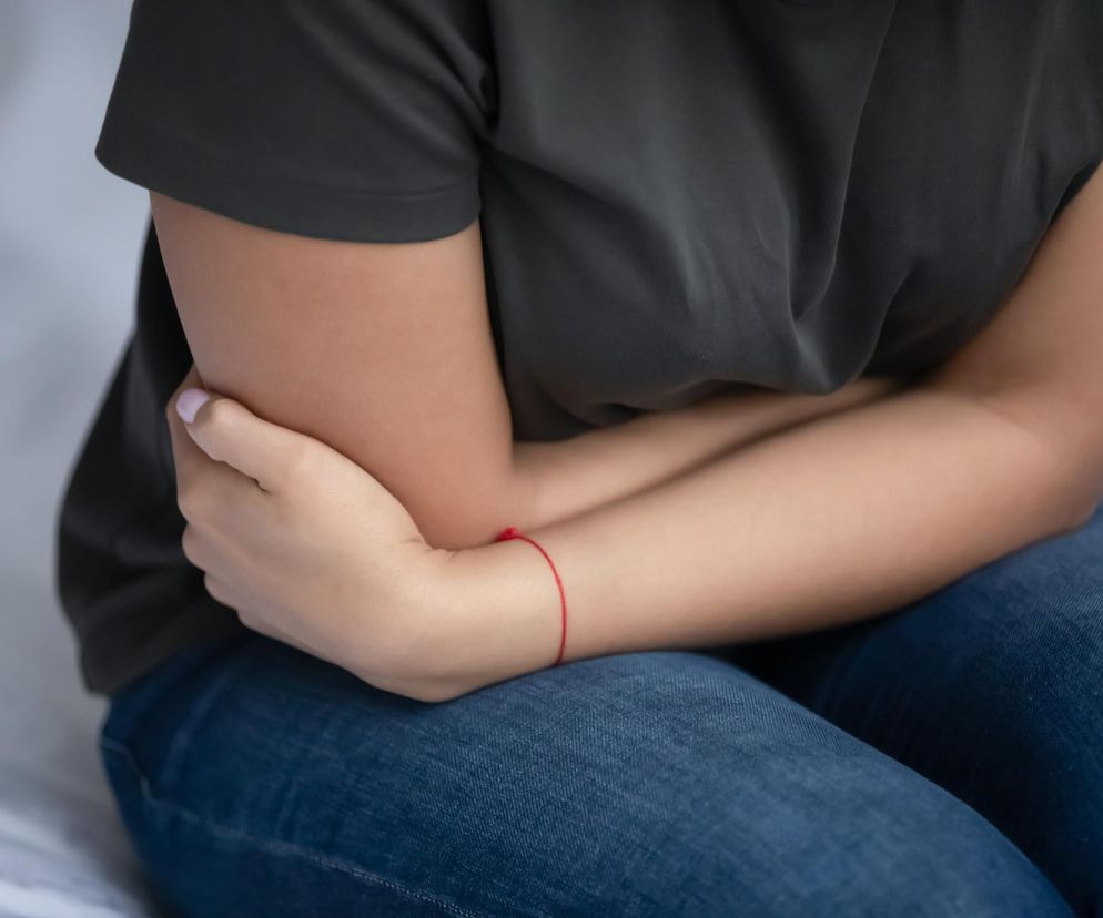 Kobieta w ciemnej koszulce i dżinsach siedzi z rękami zaciśniętymi na bolącym brzuchu, symbolizując dolegliwości jelitowe. Temat związku antybiotyków z nieswoistymi zapaleniami jelit, o którym przeczytasz na portalu Poradnik Zdrowie.