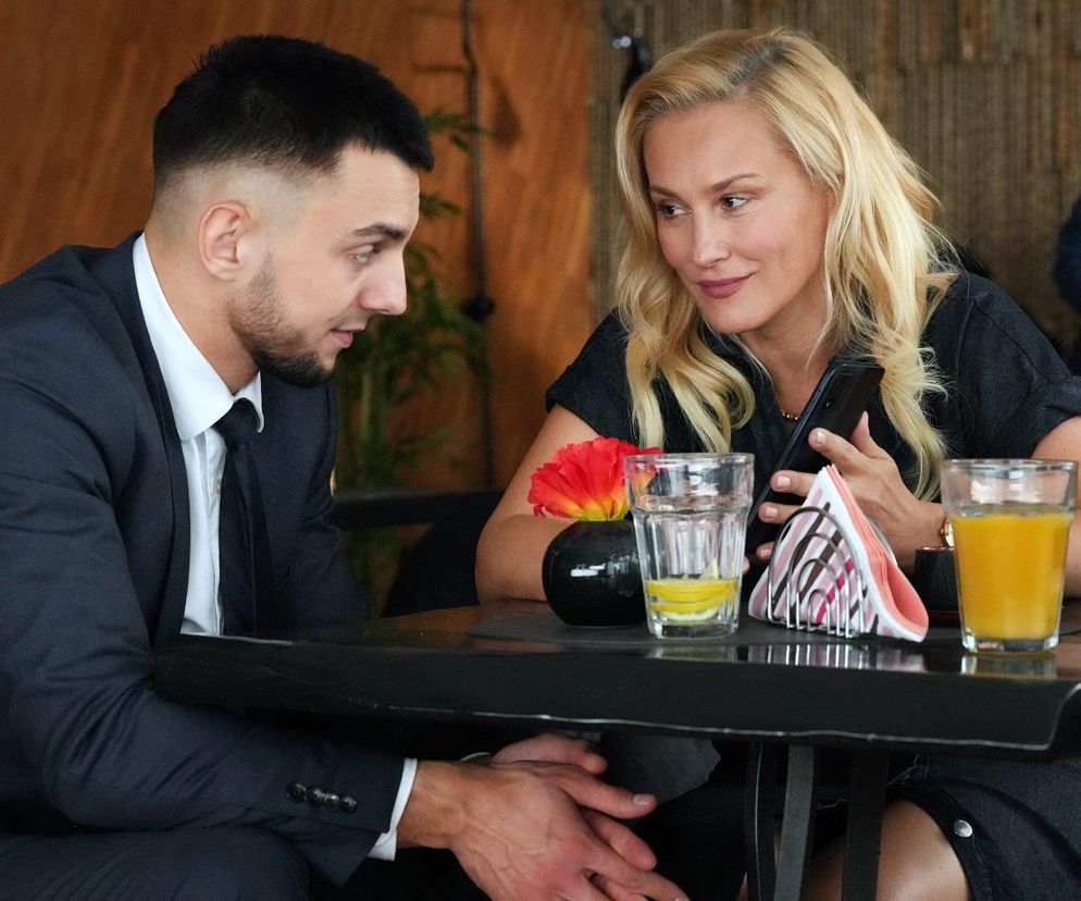 Aktorzy Oliwier Kozłowski jako Mirek i Sylwia Juszczak-Krawczyk jako Justyna Zytko siedzą przy stoliku, patrząc na siebie, co ilustruje scenę kuszenia Mirka. Blondwłosa Justyna uśmiecha się, trzymając telefon. Na stole widać napoje i serwetki. Więcej o ich losach na Super Seriale.