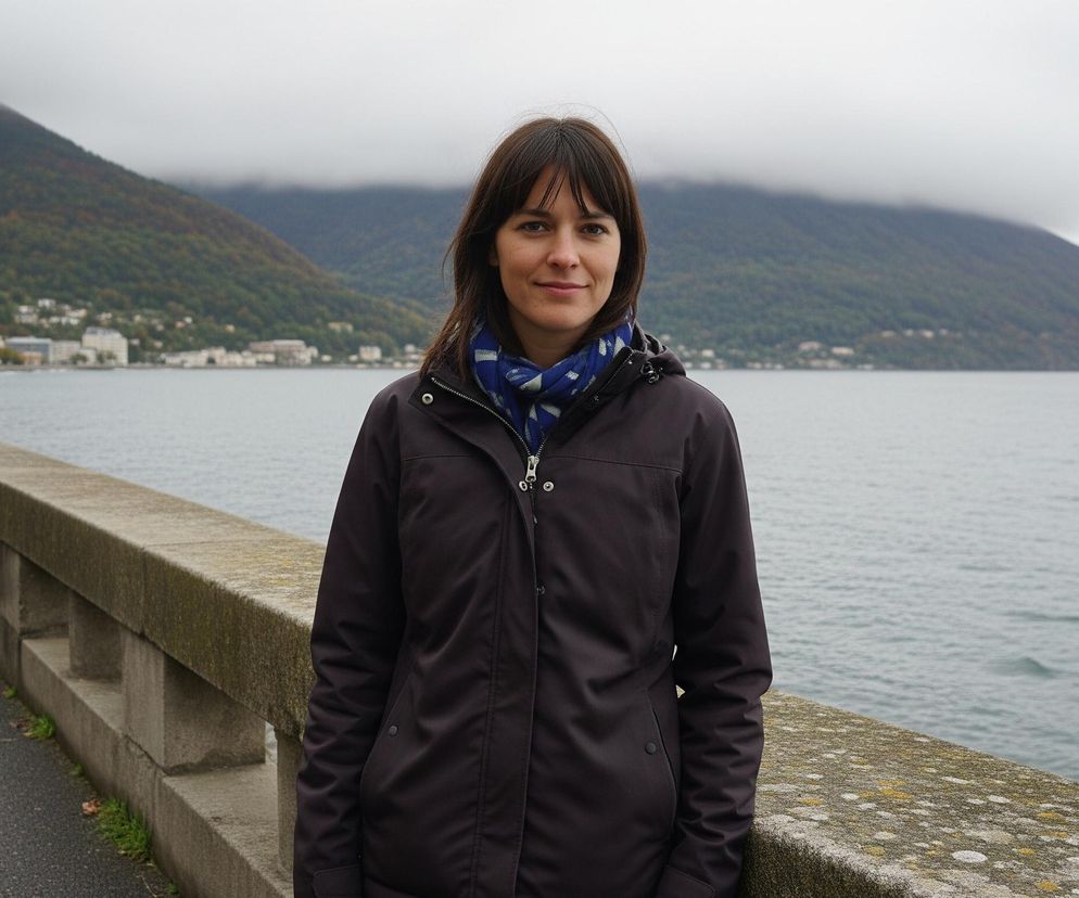 Młoda kobieta z ciemnymi włosami i uśmiechem stoi przodem do widza, ubrana w ciemną kurtkę i niebiesko-biały szalik, na tle pochmurnego nieba, morza i zalesionych gór. Znajduje się na chodniku wzdłuż betonowego muru oporowego, który oddziela go od spokojnych, szaro-niebieskich wód zatoki. Na lewym skraju widoczne są budynki miejskie u podnóża góry, natomiast zbocza pokryte są jesienną roślinnością, a ich szczyty są spowite niskimi chmurami.