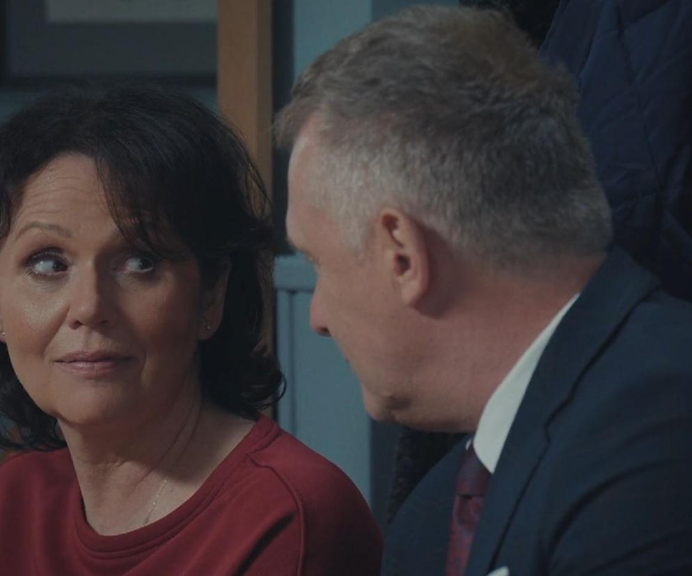 Marysia (Małgorzata Pieńkowska) w czerwonej bluzce spogląda z niepokojem na Artura Rogowskiego (Robert Moskwa) w garniturze. Scena z serialu M jak miłość podkreśla jej obawy o relację z mężem, co można przeczytać na Super Seriale.