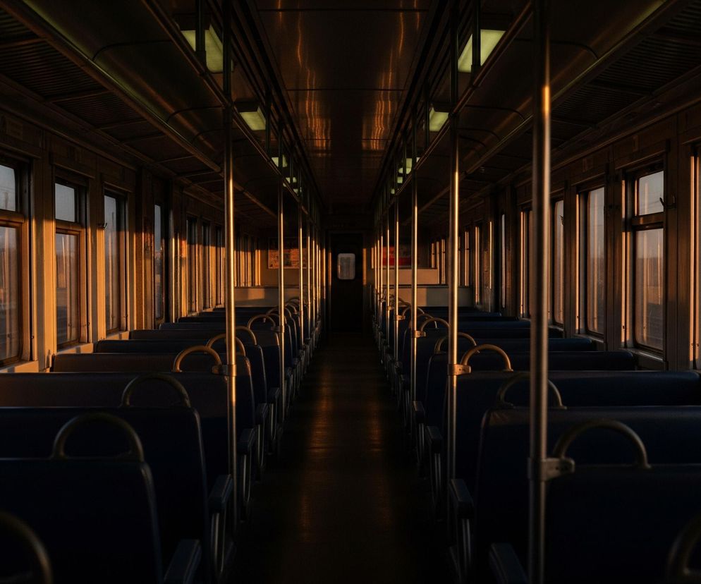 Wnętrze wagonu kolejowego rozciąga się w perspektywie, z rzędami pustych, ciemnoniebieskich siedzeń po obu stronach centralnego przejścia. Zachodzące słońce wpada przez okna po lewej stronie, rzucając długie, ciepłe, pomarańczowe pasma światła na ściany, podłogę i pionowe metalowe słupki, kontrastując z ciemnymi obszarami. Na suficie widać rzędy świetlówek, niektóre włączone, emitujące delikatne, zielonkawe światło, a na zewnątrz, przez okna, widać fragment torów i elementy krajobrazu.