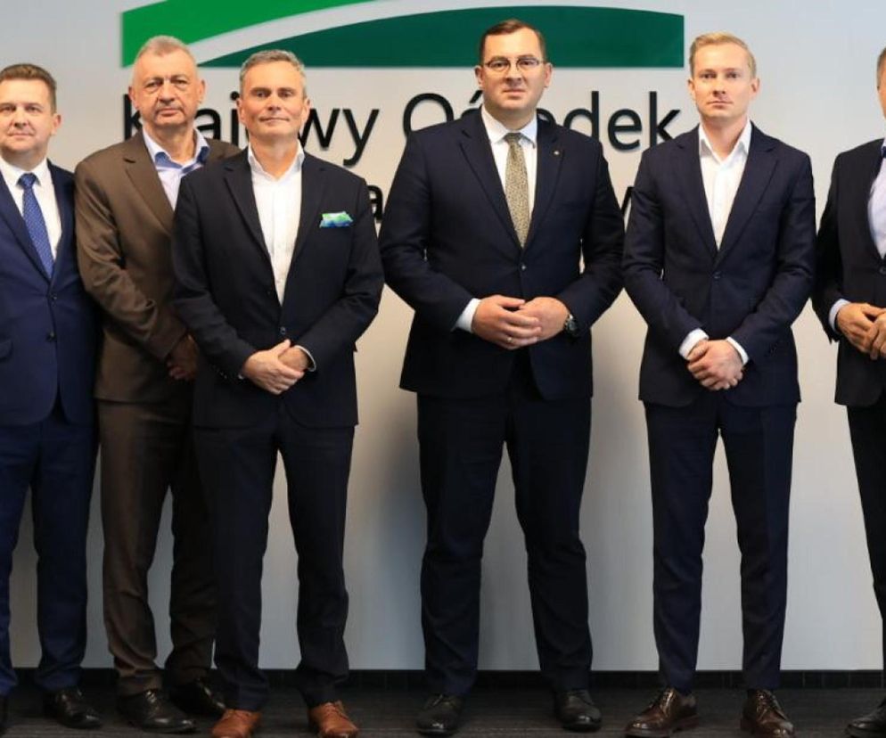 Grupa dziewięciu mężczyzn w garniturach, stojąca w siedzibie Krajowego Ośrodka Wsparcia Rolnictwa, symbolizuje porozumienie w sprawie działki pod CPK. Więcej szczegółów na temat tej sprawy można przeczytać na portalu Super Biznes.