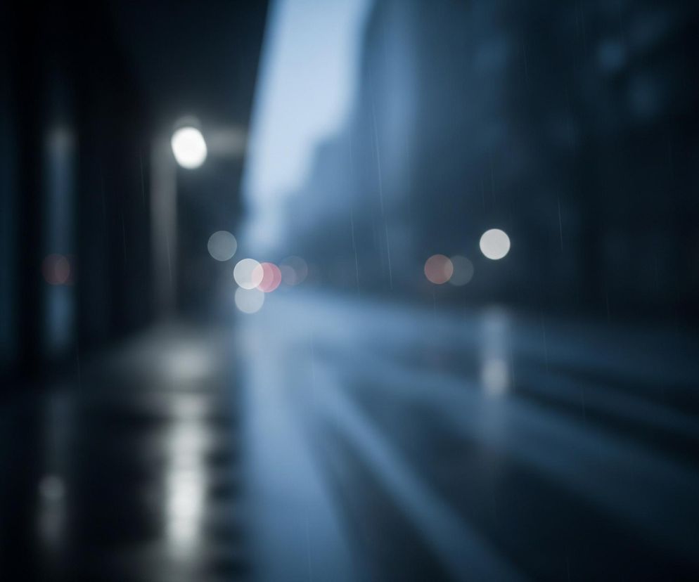 Rozmyty obraz przedstawia nocną, deszczową ulicę. Po lewej stronie widać chodnik z odbiciami świateł na mokrej nawierzchni, a nad nim zarys budynku. W centrum dominują jasne, rozmazane okręgi świateł ulicznych i samochodowych, głównie białych, czerwonych i pomarańczowych, rozciągające się w głąb perspektywy. Prawa strona ukazuje niewyraźne sylwetki budynków i drzew, z delikatnymi smugami deszczu widocznymi w powietrzu. Całość utrzymana jest w chłodnych, ciemnych odcieniach niebieskiego i szarości.