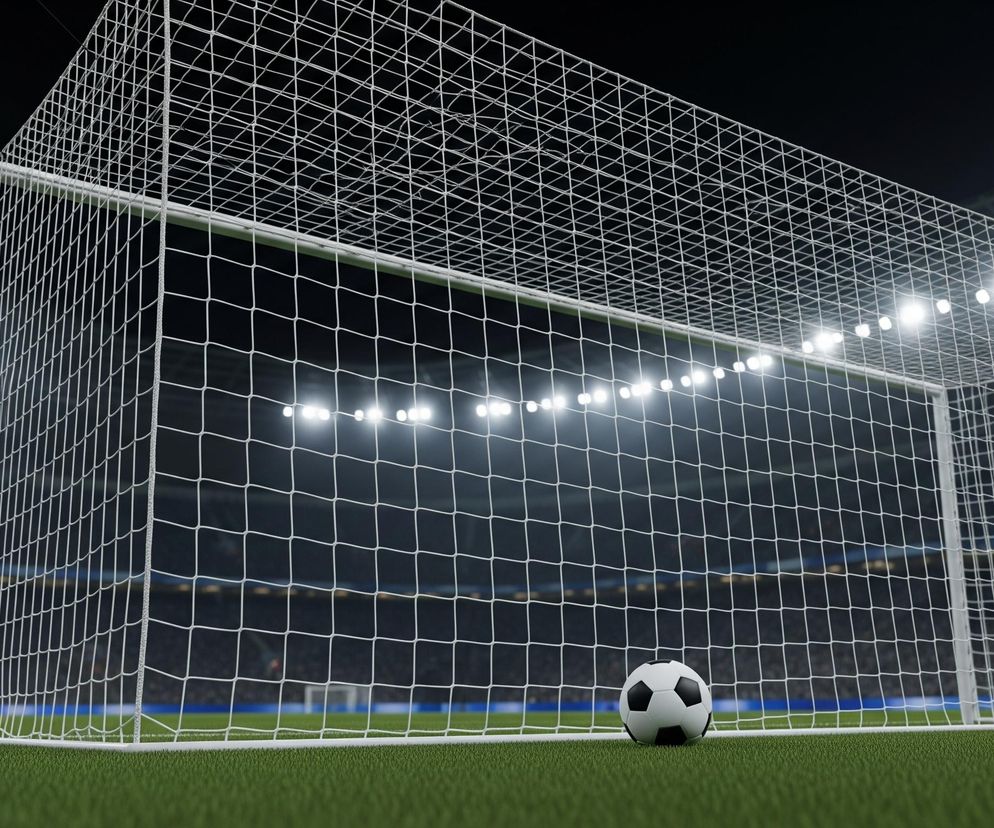 Biała bramka piłkarska z rozwieszoną siatką dominuje w centrum kadru, lekko przesunięta w prawo. Przed bramką, na zielonej trawie, leży czarno-biała piłka nożna. W tle widoczny jest ciemny stadion z rzędami siedzeń i jasnymi światłami reflektorów, oświetlającymi scenę od góry.