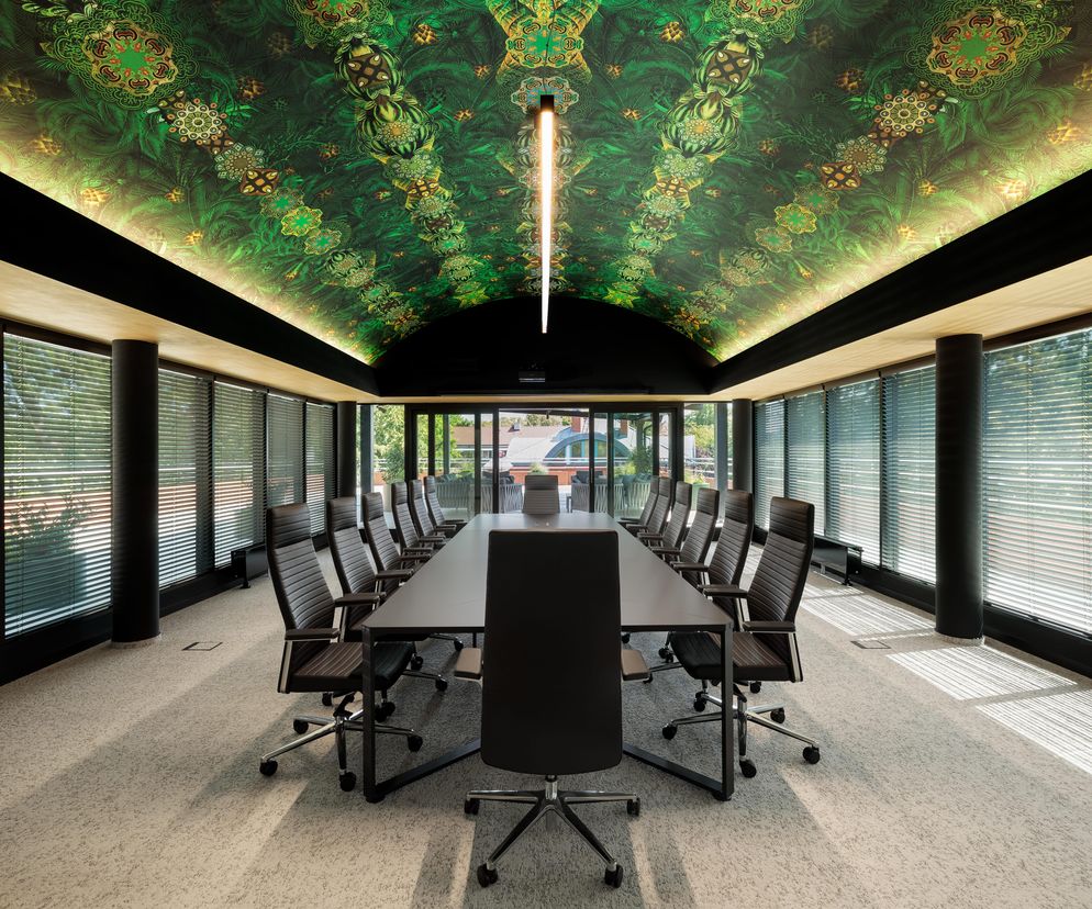Nowoczesna sala konferencyjna w siedzibie Unibaltic w Szczecinie. Charakterystyczny sufit z tapetą w zielone, tropikalne wzory z symetrycznymi, mandalami, długi stół z czarnym blatem i skórzanymi krzesłami podkreślają wyjątkowy design. Duże okna z żaluzjami zapewniają odpowiednie doświetlenie wnętrza. Więcej o modernizacji budynku Unibaltic na portalu Architektura Murator Plus.