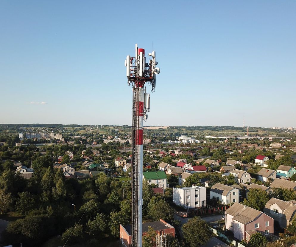 Wysoka wieża telekomunikacyjna, koloru czerwono-białego, wyposażona w liczne anteny i nadajniki, na tle miejskiego krajobrazu z licznymi domami jednorodzinnymi i zielenią. Poradnik Zdrowie informuje o technologii 5G.