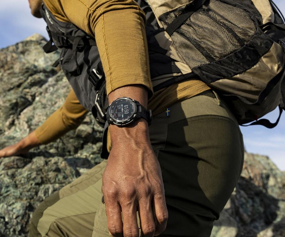 Mężczyzna w czapce z daszkiem i plecakiem wspinający się po skale, na jego nadgarstku widać smartwatch Garmin Instinct Crossover. Urządzenie łączy klasyczny styl z nowoczesnymi funkcjami, idealne dla prezesów i komandosów. Więcej o tym na Super Biznes.