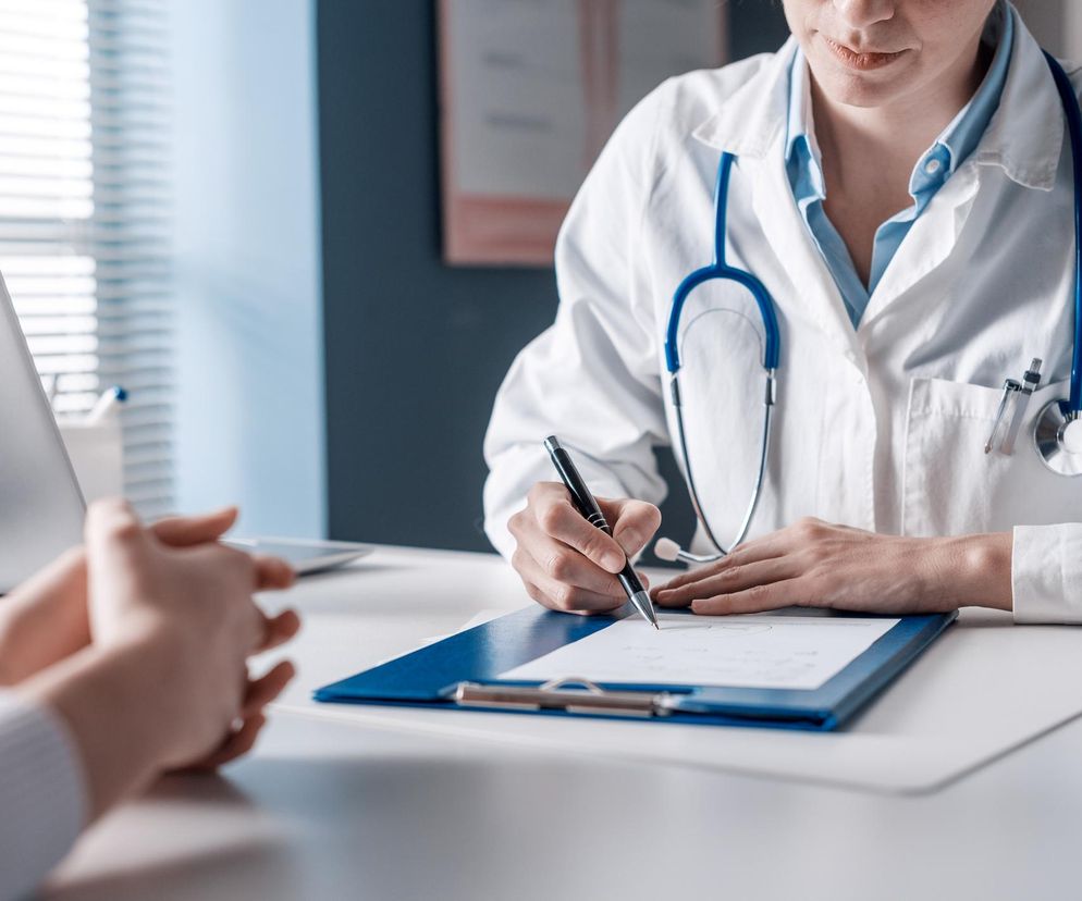 Lekarka w białym kitlu ze stetoskopem na szyi siedząca przy biurku, pisząca długopisem w karcie pacjenta. Przed nią widać dłonie pacjenta. Całość symbolizuje profesjonalizm lekarzy i ich zaangażowanie, o czym można przeczytać na portalu Poradnik Zdrowie.