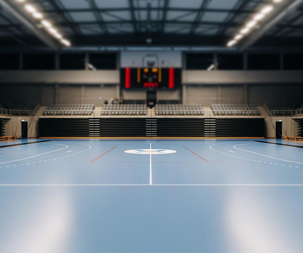 Na pierwszym planie znajduje się puste boisko sportowe o jasno niebieskiej nawierzchni z białymi i czerwonymi liniami. Po obu stronach boiska, na tle ciemnych trybun, ustawione są bramki z czerwono-białymi słupkami i białymi siatkami. W oddali widoczne są trybuny z szarymi siedzeniami oraz duży czerwono-czarny zegar cyfrowy z żółtymi i czerwonymi cyframi umieszczony na ścianie. Cała hala jest oświetlona rzędem jasnych lamp sufitowych.