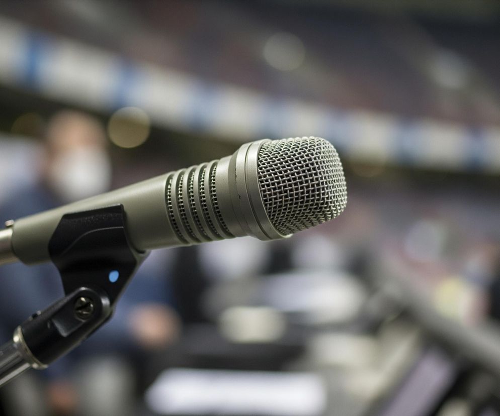 Centralnym punktem obrazu jest szary mikrofon studyjny, ustawiony na pierwszym planie po lewej stronie, skierowany w prawo. Mikrofon posiada siateczkową osłonę w górnej części oraz cylindryczny, żebrowany korpus. Jest zamocowany na czarnym, regulowanym statywie, z którego wychodzi czarny kabel. Tło jest rozmyte, ukazując niewyraźne kształty siedzeń stadionu w odcieniach niebieskiego i białego, a także zamazane sylwetki ludzi, z których jedna ma na sobie białą maseczkę. Całość obrazu utrzymana jest w stonowanej kolorystyce szarości, czerni i rozmytych barw tła.