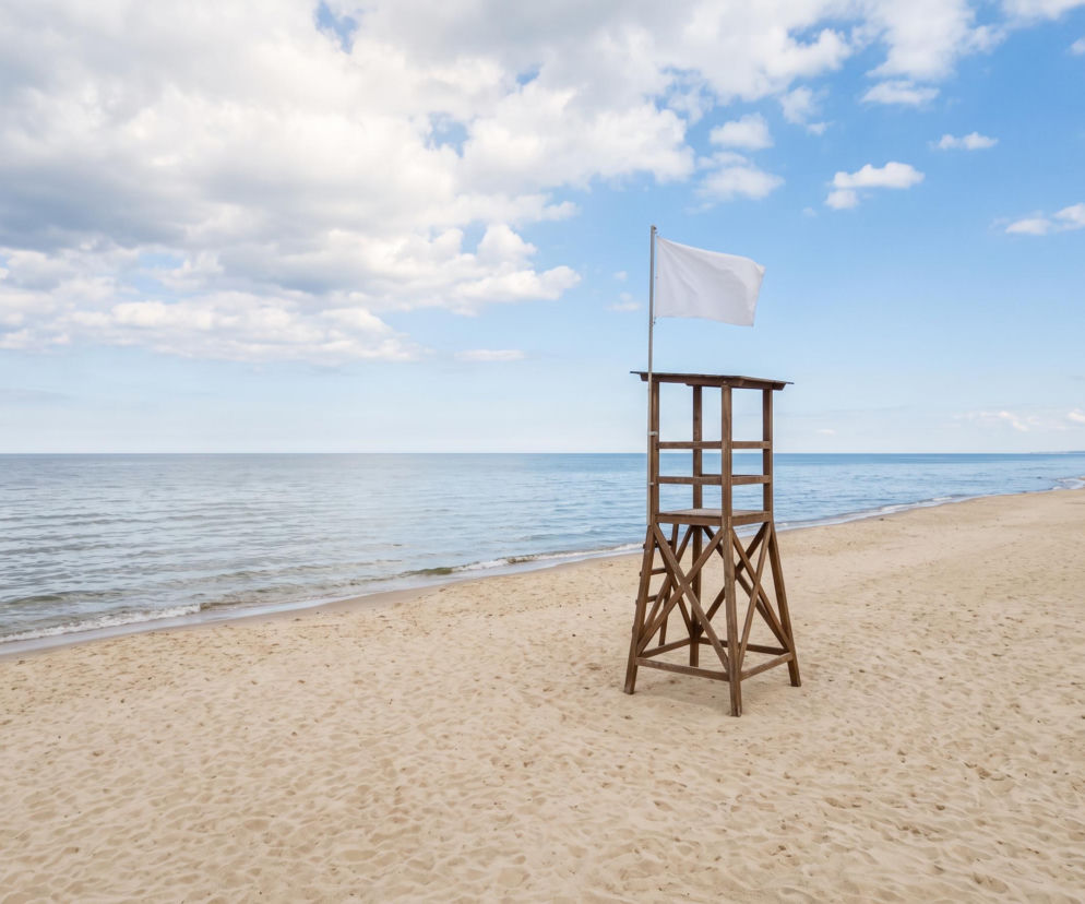 Pusta piaszczysta plaża z drewnianą wieżą ratowniczą i białą flagą. O bezpieczeństwie w Mielnie przeczytasz na SE Szczecin.