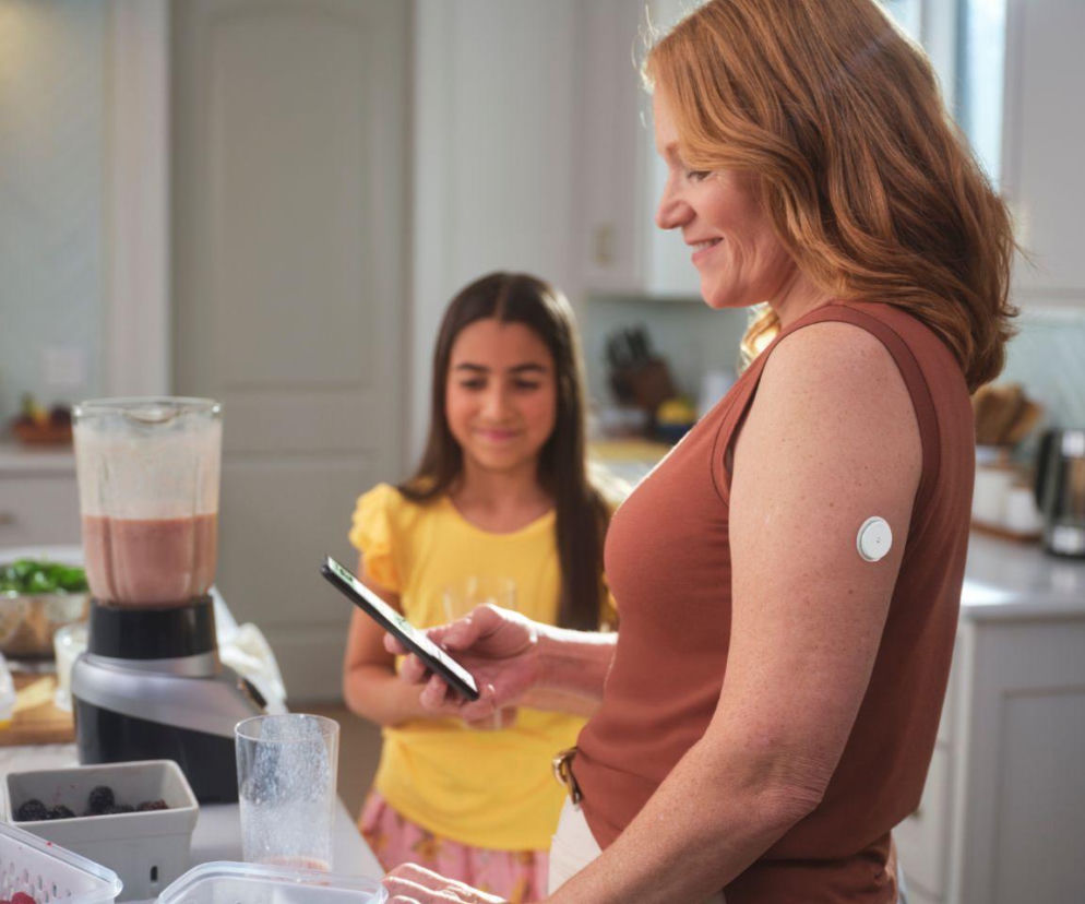 Kobieta z sensorem do ciągłego monitorowania glukozy na ramieniu, obsługująca telefon, obok dziewczynka. Obie są w kuchni, a na blacie widać blender i owoce. Dzięki nowoczesnym systemom monitorowania glukozy, o których przeczytasz na Poradnik Zdrowie, życie z cukrzycą staje się bezpieczniejsze i spokojniejsze.