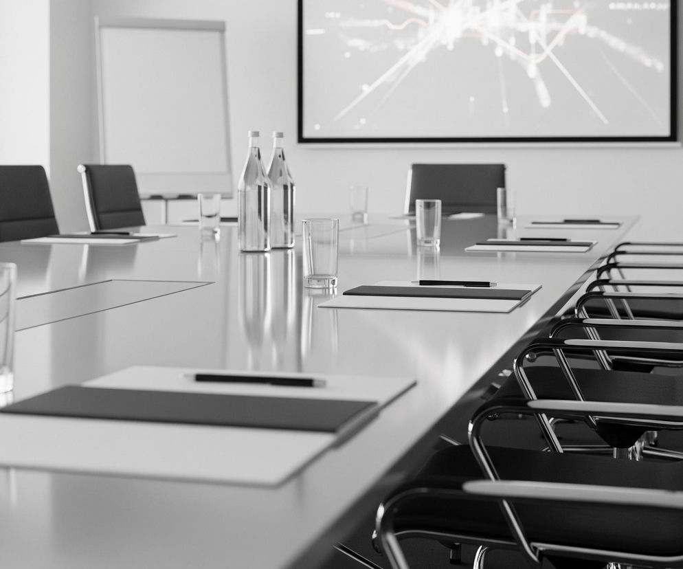 Długi, prostokątny stół konferencyjny z lśniącym, jasnoszarym blatem dominuje w pomieszczeniu, odbijając światło. Wzdłuż stołu ustawiono szereg krzeseł z ciemnymi, prawdopodobnie skórzanymi siedziskami i oparciami, oraz błyszczącymi, metalowymi ramami, z których wiele jest widocznych w zbliżeniu po prawej stronie. Na stole rozłożone są białe notatniki z czarnymi, wąskimi podkładkami i długopisami, a także szklanki z wodą i dwie butelki wody w przezroczystego szkła, stojące bliżej końca stołu. Na przeciwległej ścianie wisi ekran projekcyjny z wyświetlonym abstrakcyjnym wzorem jasnych linii na ciemniejszym tle, a obok niego widoczna jest również biała tablica.