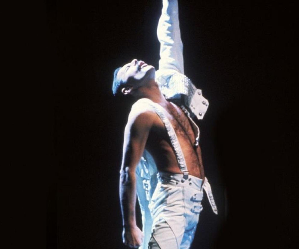 Freddie Mercury w białym stroju scenicznym z podniesioną ręką. O darmowej wystawie w Londynie przeczytasz na EskaRock.