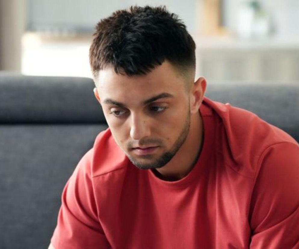 Mirek (Oliwier Kozłowski) z serialu Barwy szczęścia w czerwonej koszulce, zaniepokojony po oblaniu matury, siedzi na szarej kanapie. Jego twarz wyraża smutek i bezsilność. Więcej o losach bohatera znajdziesz na Super Seriale.