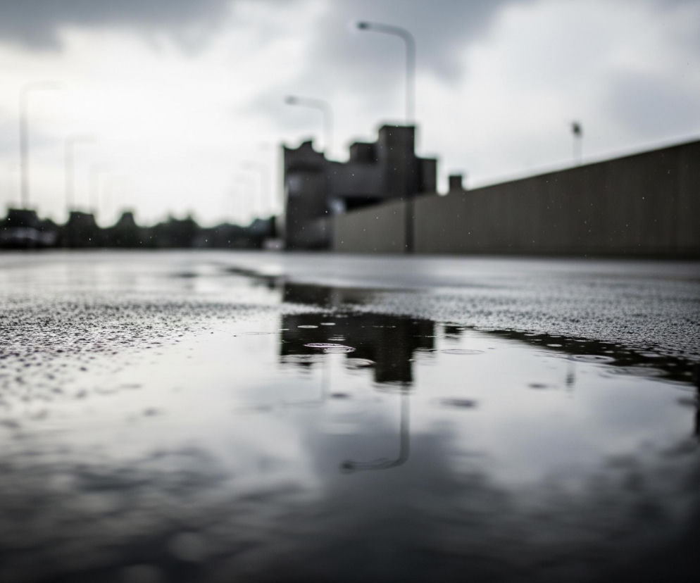 Szeroka kałuża wody pokrywa większość pierwszego planu, odbijając pochmurne, szare niebo i sylwetki odległych słupów. Na powierzchni kałuży widać liczne, drobne kręgi tworzone przez spadające krople deszczu, niektóre z nich są zamazane, co sugeruje ruch. W tle widoczna jest rozmyta infrastruktura miejska: zarysy budynków i latarni ulicznych w odcieniach szarości, z których jeden wysoki słup rozciąga się pionowo w lewym centrum, a inny po prawej stronie. Ogólny nastrój obrazu jest ponury i deszczowy, z dominacją ciemnych, chłodnych tonów.