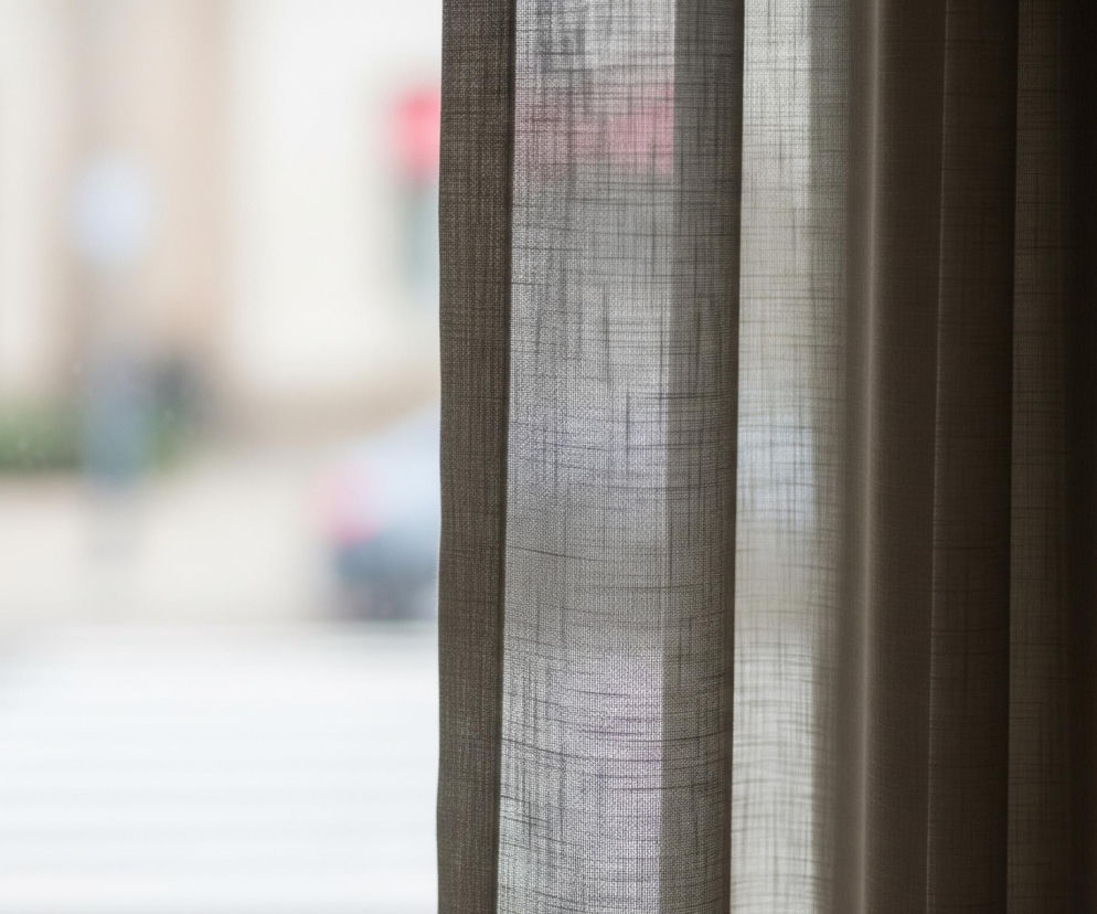 Po prawej stronie obrazu widać pionowe, teksturowane zasłony w odcieniach szarości i brązu, częściowo zaciemnione, z widocznym przez nie światłem, które ukazuje subtelną siatkę tkaniny. Po lewej stronie, za zasłonami, rozciąga się rozmyty widok ulicy z białymi pasami przejścia dla pieszych na szarym asfalcie, a dalej w tle, niewyraźne kształty budynków, drzew i prawdopodobnie pojazdu, częściowo z czerwonym akcentem. Całość tworzy kontrast między ostrością i teksturą zasłon a miękkim, rozmytym tłem.