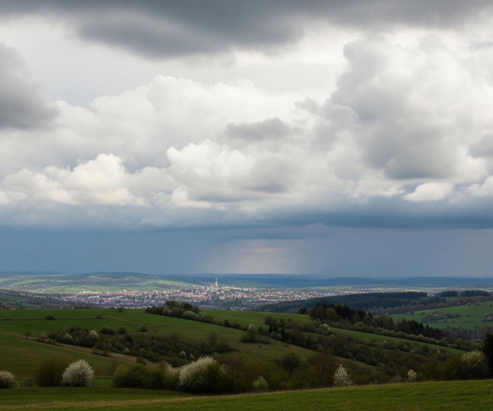 Rozległy krajobraz charakteryzuje się pofałdowanymi zielonymi wzgórzami z rzędami drzew i krzewów kwitnących na biało w dolnej części kadru. Pośrodku obrazu, w oddali, rozciąga się miejska zabudowa z licznymi budynkami i wyraźnie widocznymi wieżami kościelnymi. Nad horyzontem dominują gęste, szare chmury, a po prawej stronie widać rozświetlony promień słońca, wskazujący na przejaśnienie lub padający deszcz.