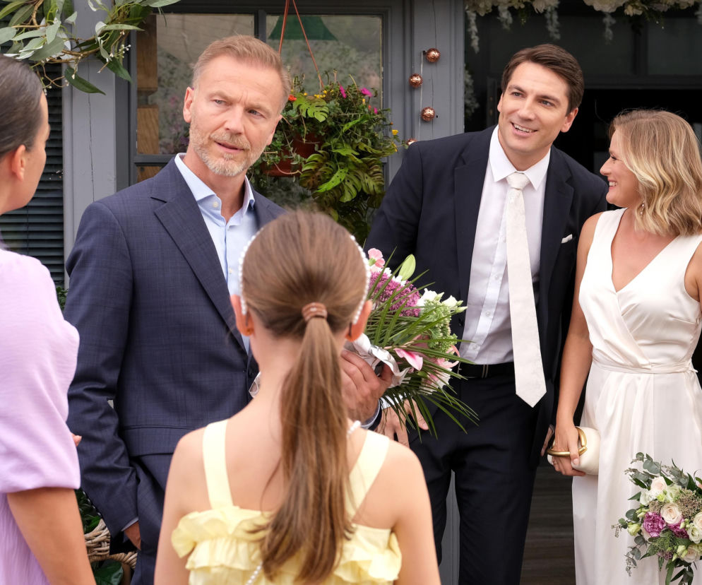 Julia (Marta Chodorowska) w jasnej sukience z dekoltem kopertowym i bukietem ślubnym stoi obok Dima (Michaił Pszeniczny) w ciemnym garniturze i jasnym krawacie, oboje uśmiechnięci. Obok nich stoją Magda (Anna Mucha) w liliowej sukience i Andrzej (Krystian Wieczorek) w garniturze. Na pierwszym planie Nadia (Mira Fareniuk) w żółtej sukience, tyłem do obiektywu. Scena ze ślubu Julii i Dimy w serialu „M jak miłość”, o czym przeczytasz więcej na portalu Super Seriale.