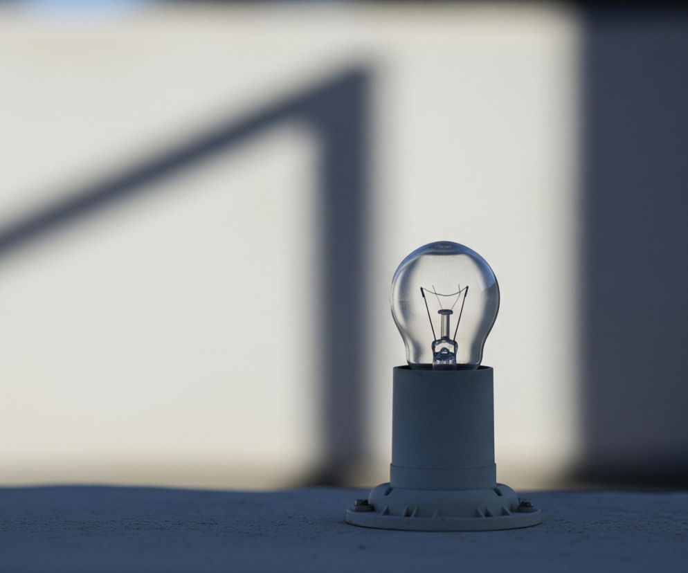 Przezroczysta żarówka z cienkim, spiralnym żarnikiem w środku, osadzona jest w białej, walcowatej oprawce na pierwszym planie obrazu. Oprawka ma szeroką, okrągłą podstawę z trzema widocznymi śrubami mocującymi. Obiekt stoi na ciemnoszarym, lekko chropowatym podłożu, które rozciąga się wzdłuż dolnej części kadru. Tło stanowi jasna, beżowo-biała powierzchnia z wyraźnymi, ciemnoszarymi cieniami w kształcie geometrycznych linii, biegnącymi ukośnie i pionowo. Górna część tła wydaje się być delikatnie rozmyta i jaśniejsza.