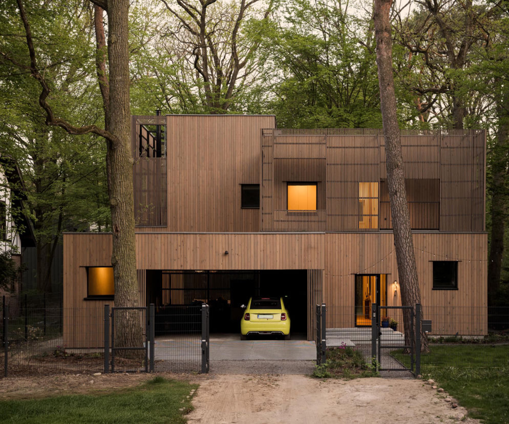 Nowoczesny dom S31 w Podkowie Leśnej, zbudowany z drewnianych paneli, harmonijnie wtapia się w otoczenie starodrzewu. Architektura Murator Plus opisuje takie projekty, które szanują naturę i lokalne regulacje konserwatorskie. Jasno świecące okna kontrastują z zielonym tłem.