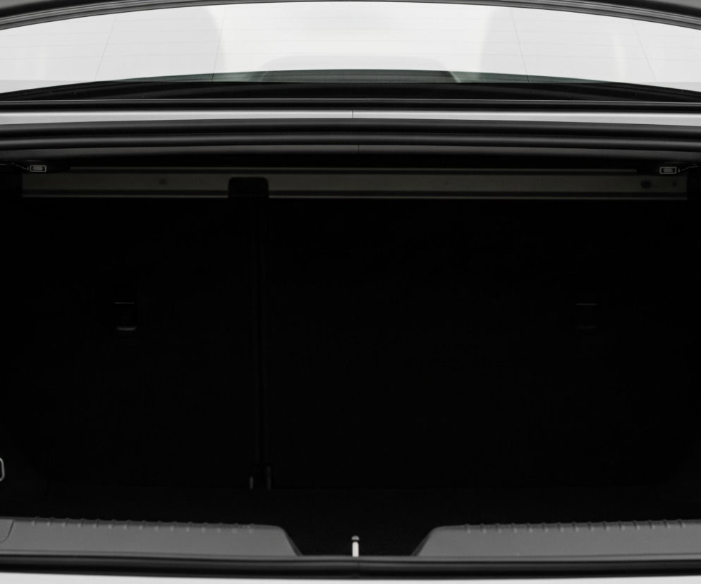 Otwarty bagażnik samochodu, z perspektywy patrzącego bezpośrednio do środka. Wnętrze bagażnika, w większości pokryte ciemnym materiałem, jest puste. Górna część bagażnika to jasnoszara klapa, od której odchodzi karbowana, czarna rurka z lewej strony oraz mechanizm zawiasu z prawej. Na dnie bagażnika znajdują się dwa metalowe uchwyty w kształcie litery U, po jednym z każdej strony.