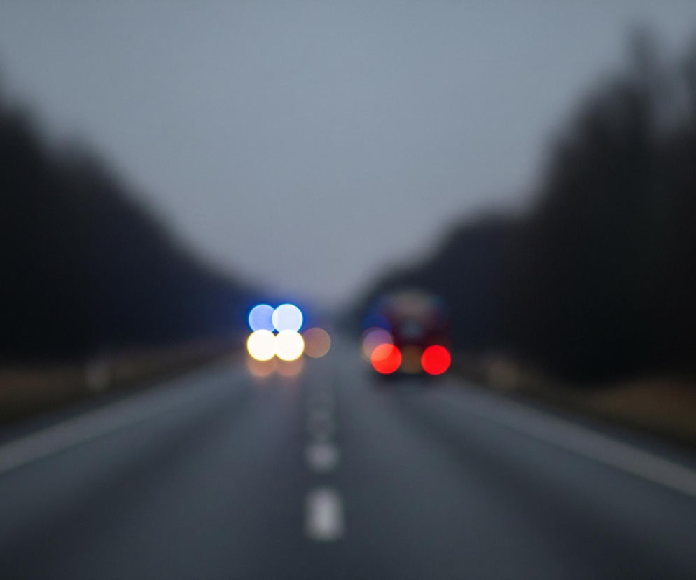 Rozmazany widok drogi asfaltowej w dzień o szarej, pochmurnej aurze. Na środku drogi widoczne są przerywane białe linie, z kolei linie ciągłe oddzielające pasy ruchu są po prawej i lewej stronie. W oddali, na horyzoncie, rozmyte drzewa tworzą ciemne sylwetki po obu stronach drogi, zaś niebo jest jasnoszare. Na drodze, w oddali, znajdują się dwa rozmazane pojazdy z włączonymi światłami: jeden ma światła niebieskie i żółte, a drugi czerwone.