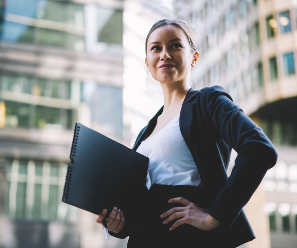 Uśmiechnięta bizneswoman z teczką na tle biurowców. O zbliżającym się Forum Inwestorek przeczytasz na Eska.