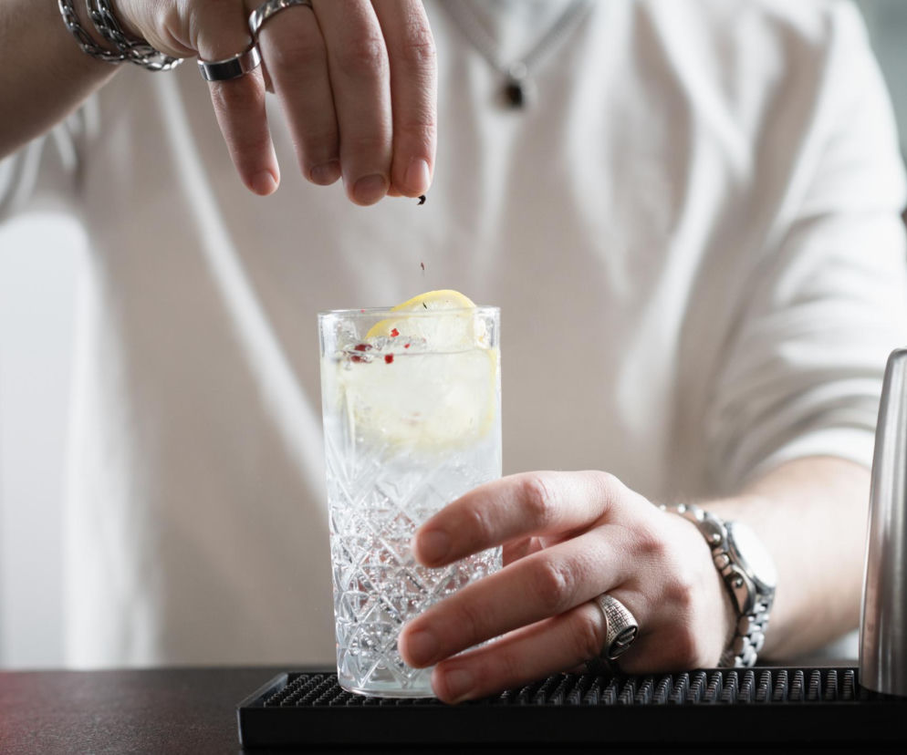 Osoba w białej koszulce dodaje suszone zioła do szklanki ze stacked water z plastrem cytryny i lodem. Mężczyzna ma srebrne pierścionki i bransoletkę, co podkreśla nowoczesny trend. Więcej o stacked water przeczytasz na portalu Poradnik Zdrowie.