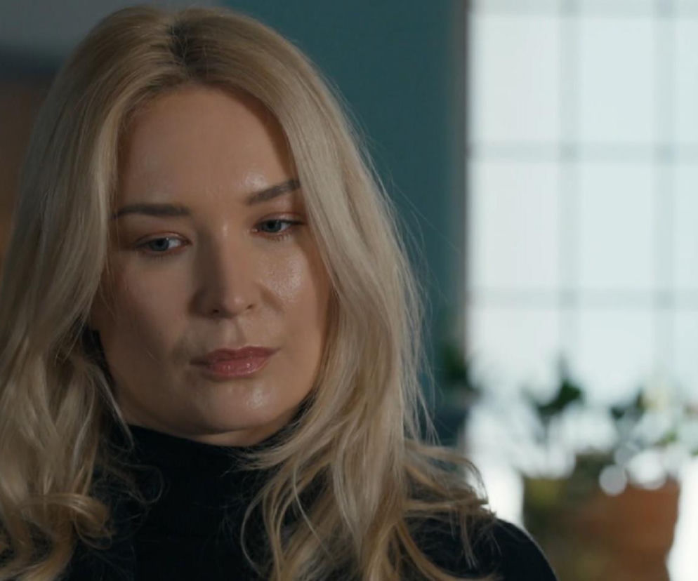 Zbliżenie na Martę Dąbrowską, aktorkę wcielającą się w Karolinę z Barw szczęścia. Blondynka o niebieskich oczach patrzy w dół z zamyśleniem, jej usta są lekko rozchylone. Nosi czarny golf, w tle niewyraźnie widać wnętrze. Sprawdź, co dalej z jej bohaterką na Super Seriale.