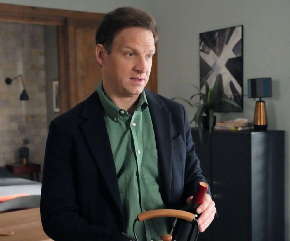 Michał Rolnicki jako Łukasz Sadowski z Barw szczęścia w zielonej koszuli i granatowej marynarce, trzymający butelkę wina z korkociągiem. Aktor w mieszkaniu z dekoracjami i sypialnią w tle. Więcej o losach Łukasza na Super Seriale.