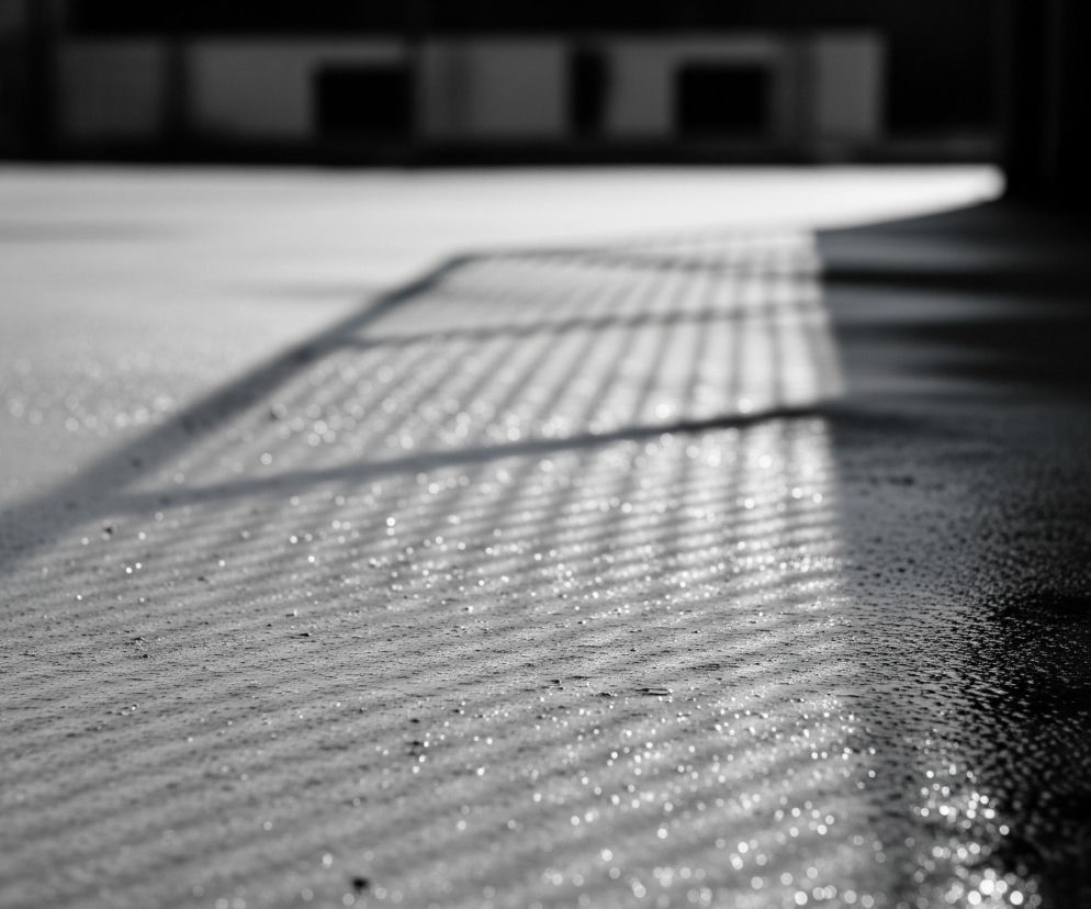 Szara, mokra powierzchnia zajmuje większą część obrazu, pokryta drobnymi kroplami wody, które odbijają światło, tworząc efekt bokeh. Na tej powierzchni widoczny jest wzór regularnych, równoległych cieni, przecinających ukośnie prawą stronę kadru. W górnej części zdjęcia, poza ostrością, znajduje się rozmyte tło, przedstawiające ciemne, geometryczne kształty budynków lub konstrukcji. Prawa strona kadru jest znacznie ciemniejsza, przechodząc w niemal czarny obszar bez wyraźnych szczegółów.