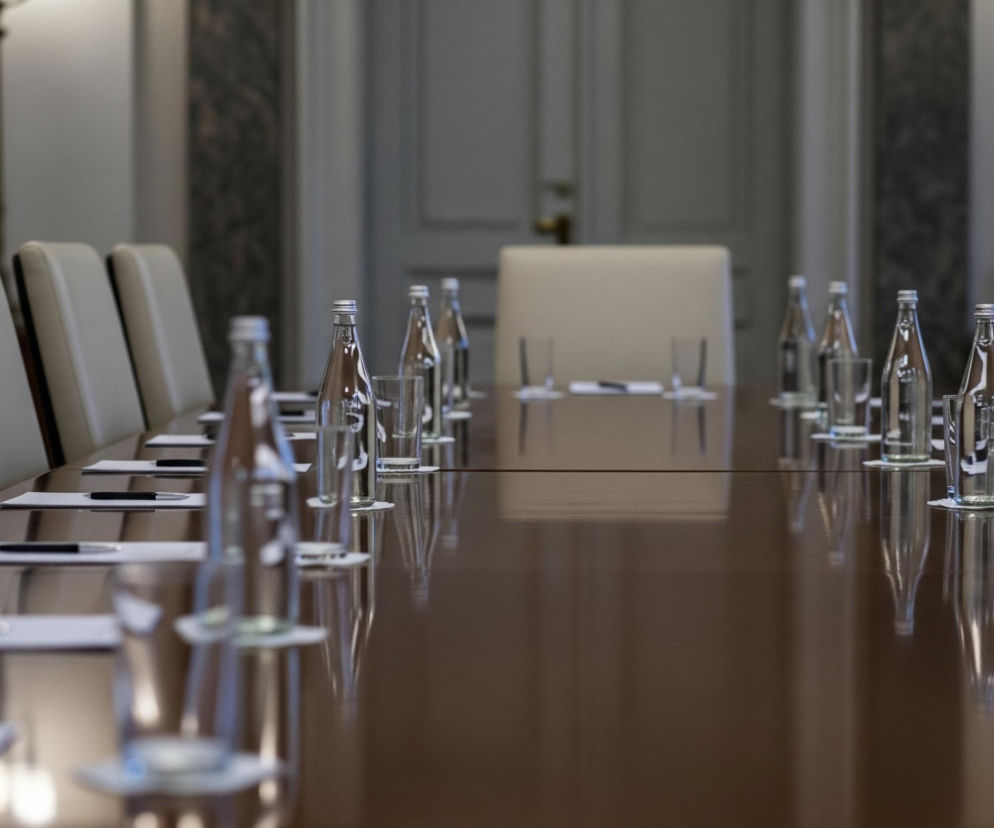 Duży, polerowany, brązowy stół konferencyjny rozciąga się przez środek kadru, odbijając światło. Po obu stronach stołu ustawione są jasne, beżowe, skórzane krzesła. Na stole wzdłuż obu krawędzi leżą białe notatniki z czarnymi długopisami, a przed każdym miejscem znajduje się butelka wody i pusta szklanka, stojące na białych podkładkach. W tle widać jasną ścianę z delikatnymi zdobieniami i ciemne framugi drzwi.